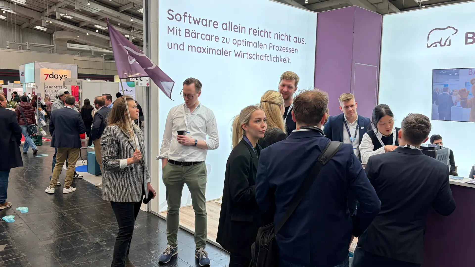 Ein gut besuchter Messestand von Bärcare auf der Pro Care 2026. Im Hintergrund ist eine große, hell leuchtende Wand mit der Aufschrift „Software allein reicht nicht aus. Mit Bärcare zu optimalen Prozessen und maximaler Wirtschaftlichkeit.“ zu sehen. Mehrere Personen in Business-Outfit stehen am Stand im Gespräch zusammen, links hält eine Frau eine lila Flagge mit dem Bärcare-Logo.