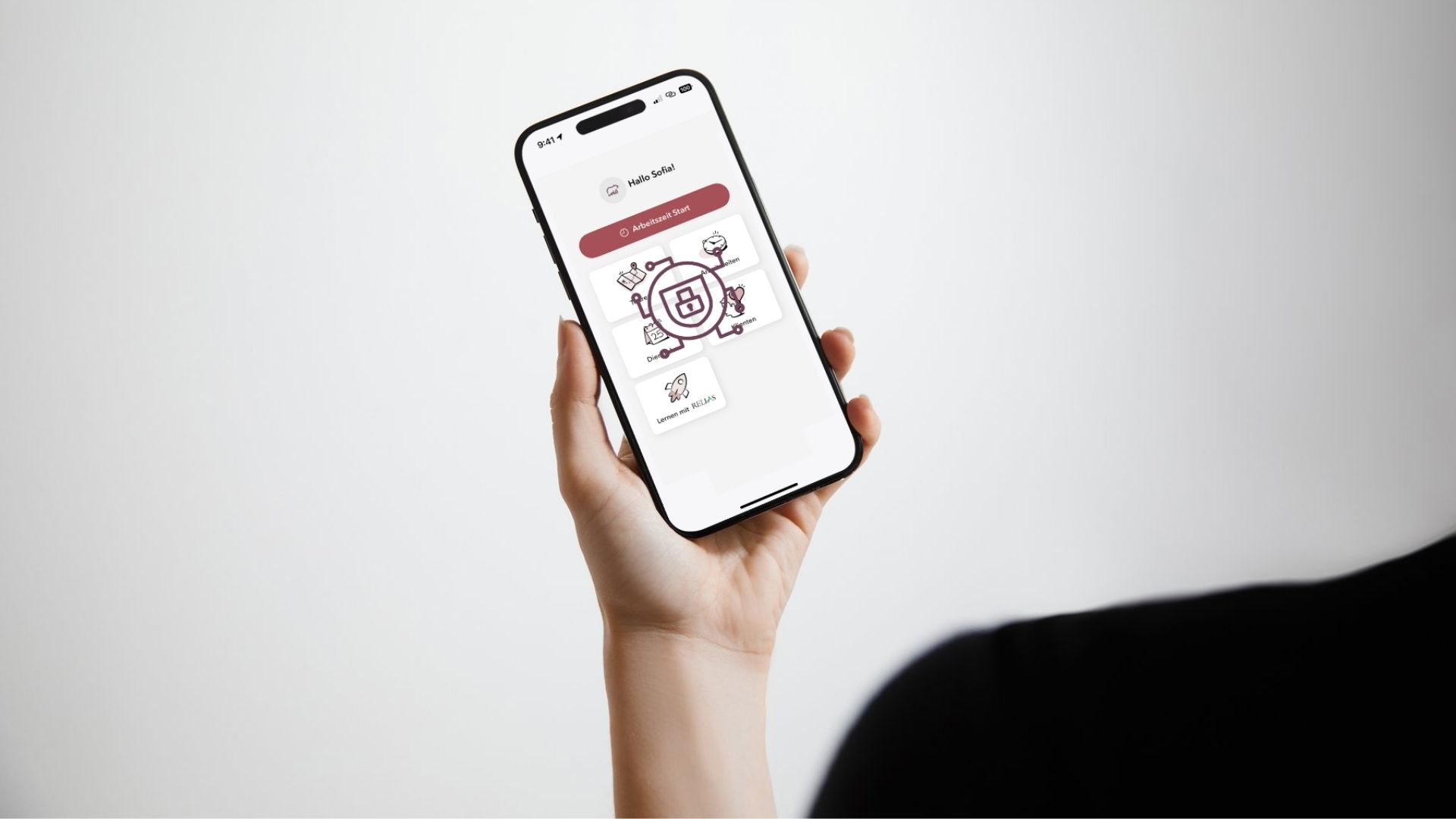 Eine Person hält ein iPhone in der Hand, auf dem eine App-Oberfläche für ambulante Pflegedienste angezeigt wird. Der Bildschirm zeigt eine Begrüßung („Hallo Sofia!“), einen großen Button „Arbeitszeit Start“ sowie mehrere Symbole mit Beschriftungen wie „Digi“, „Lernen mit RELIAS“ und „Klienten“. Im Zentrum des Displays ist ein grafisches Datenschutzsymbol mit Schloss zu sehen, das für Datensicherheit steht. Der Hintergrund ist hell und minimalistisch.
